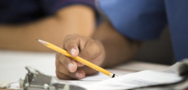 A closeup of a young man's hand holding a pencil and resting on a clipboard with papers
